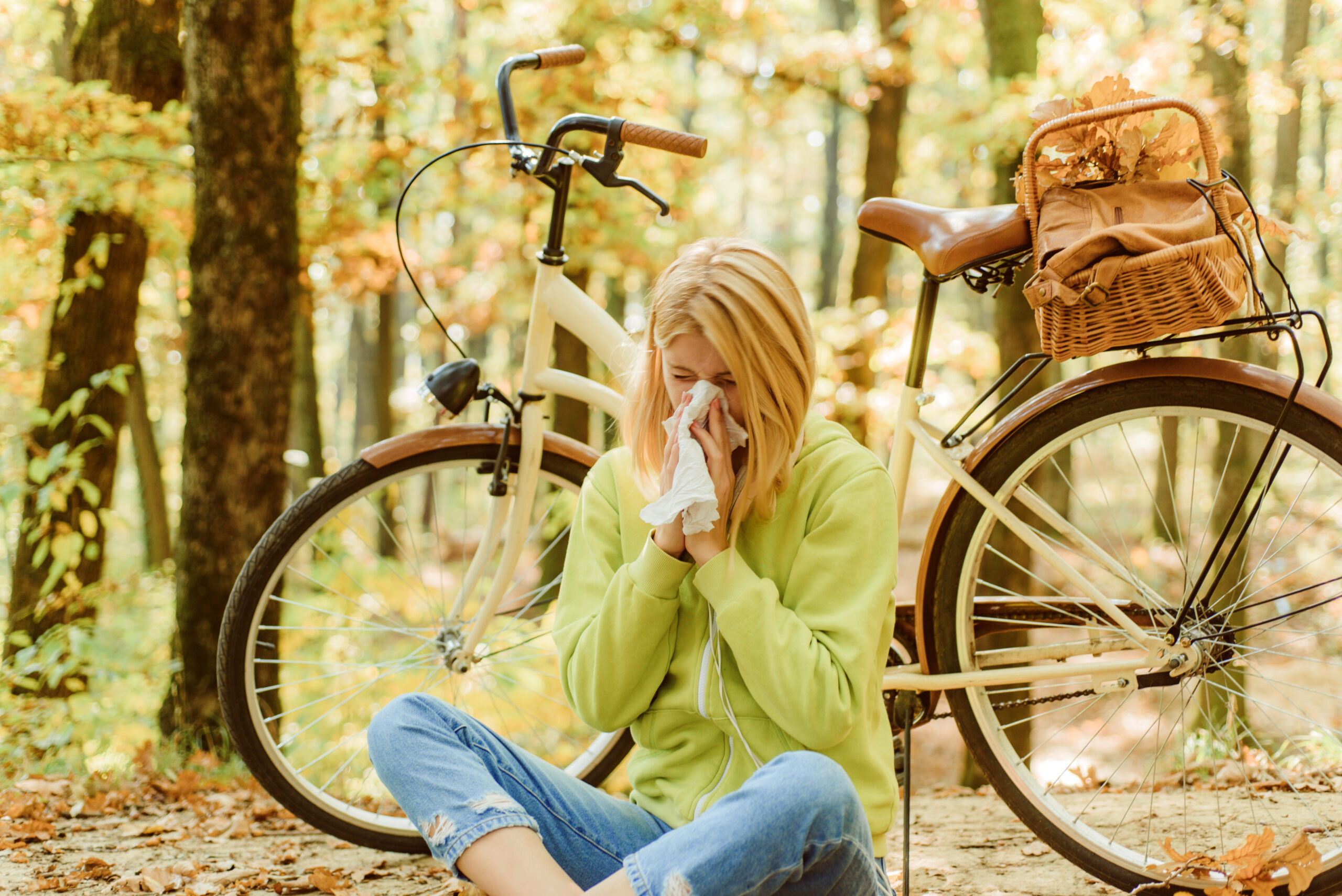 A woman taking a break from riding her bike on a trail in the woods to blow her nose as she is suffering from fall allergies.